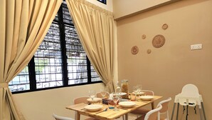 Un coin repas avec une table dressée pour quatre personnes, entourée de chaises en osier, d’une chaise haute blanche et d’une fenêtre avec des rideaux.