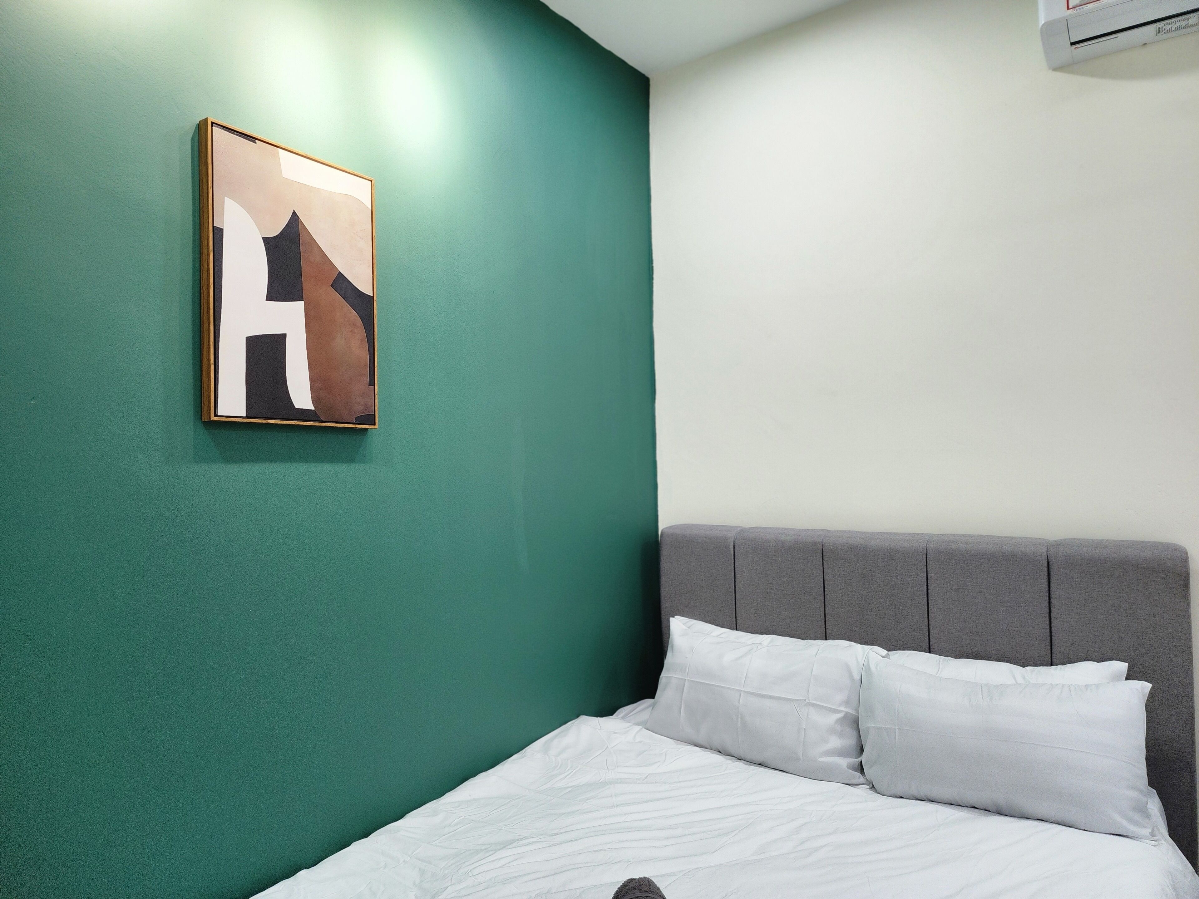 A neatly made bed with white bedding, a grey upholstered headboard, and a teal wall with a framed abstract artwork.