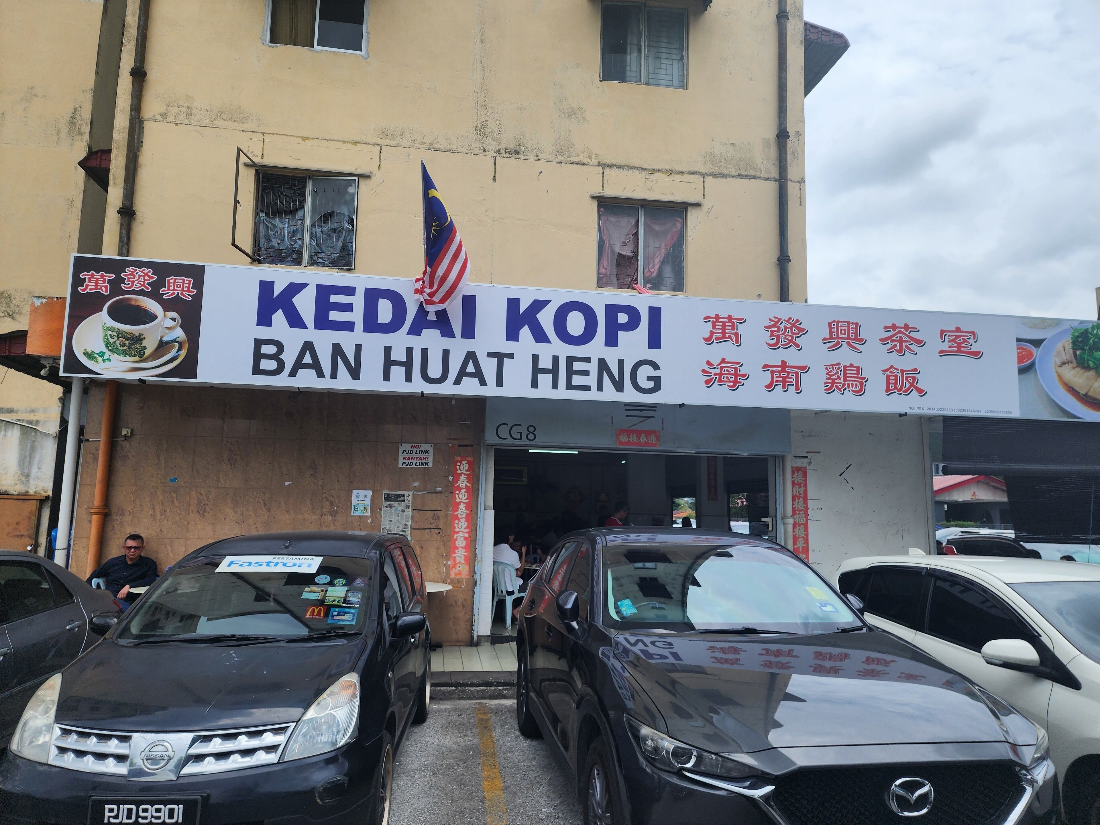 A building with a sign for 'KEDAI KOPI BAN HUAT HENG' and a Malaysian flag. There are cars parked in front.