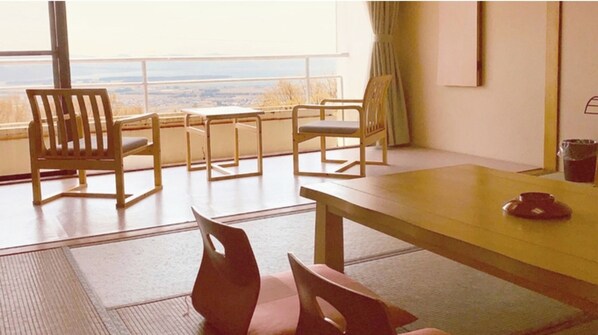 A room with a wooden table, four chairs, a view through large windows, and a trash bin.
