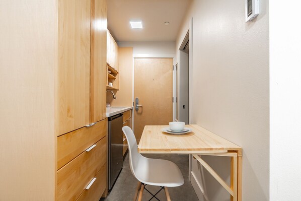 A compact kitchen with wooden cabinets, a white chair, and a small table with a bowl. - Broadway-Condotel on First Hill (Seattle)