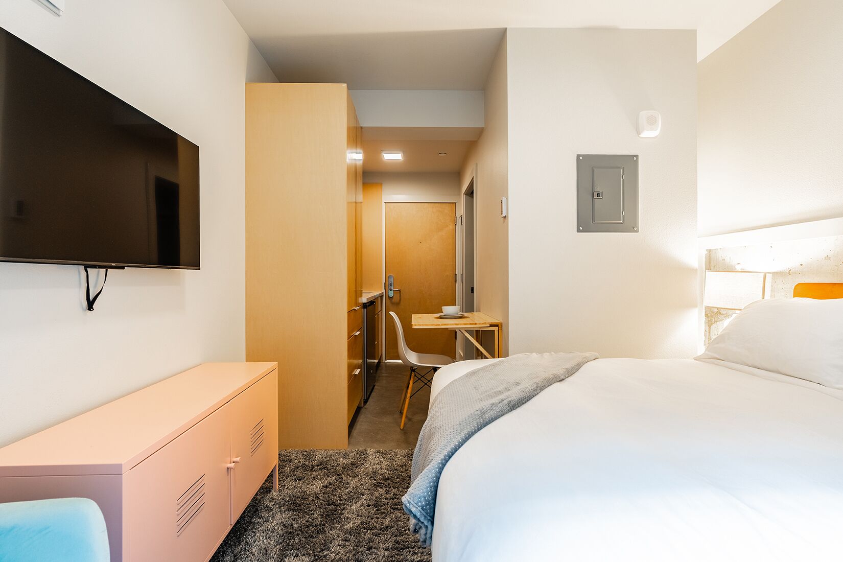 Una habitación de hotel con cama, televisor montado en la pared, escritorio con silla y armario.