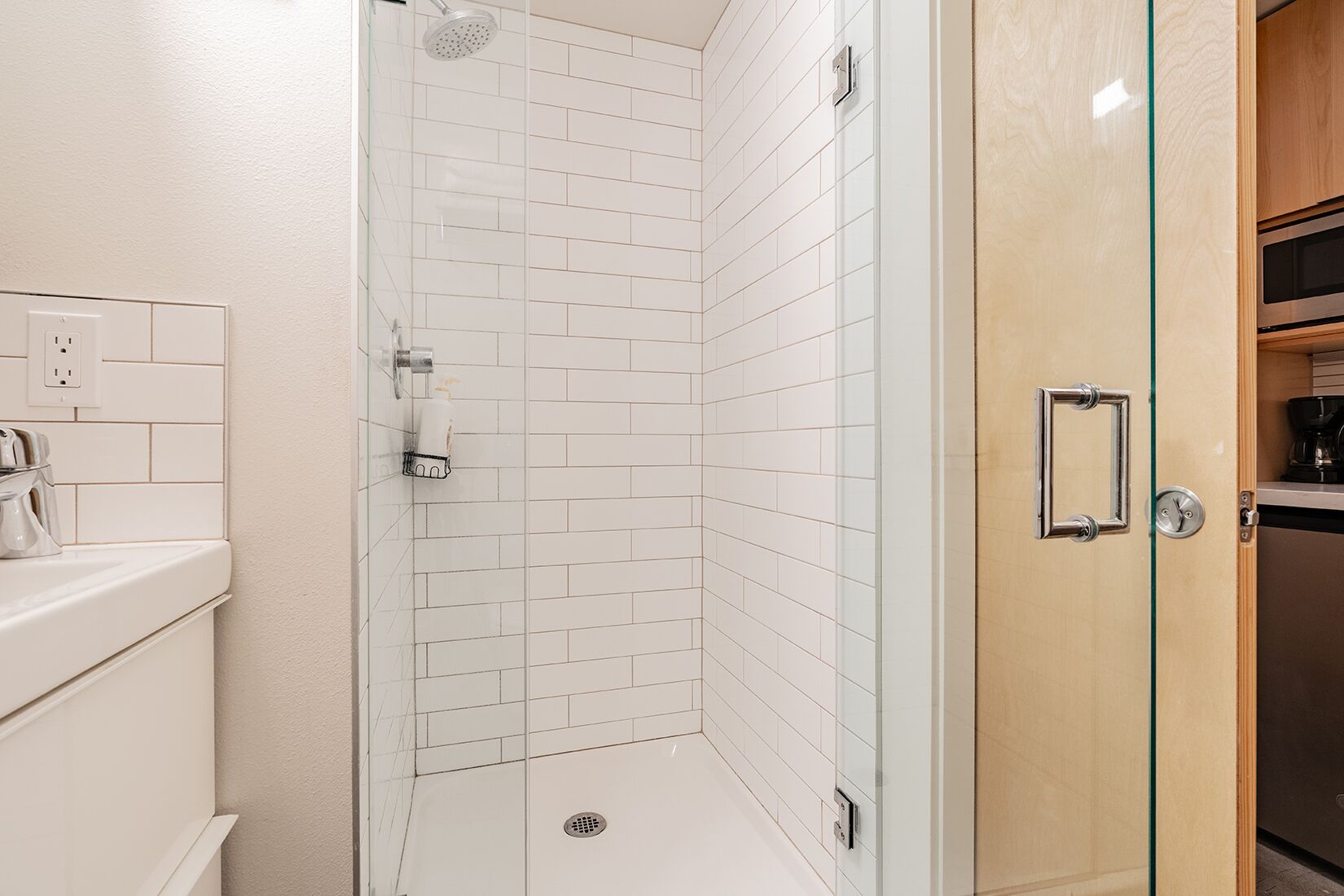Une salle de bain moderne avec des carreaux de céramique blancs de type « métro », une douche en verre, un meuble en bois avec une machine à café et des toilettes dotées d’un distributeur de savon.