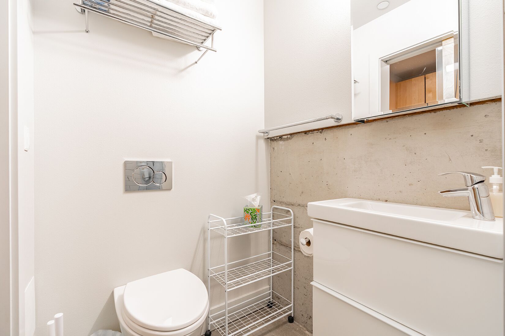 Ein kleines, sauberes Badezimmer mit einer weißen Toilette, einem Metallhandtuchhalter, einem Ablagefach mit Toilettenartikeln, einem weißen Waschbecken mit Spiegel darüber und einer Betonwand.