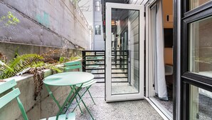 Een terras met groene tafels en stoelen, een betonnen muur met beplanting, en een ingang van een gebouw met een glazen deur.