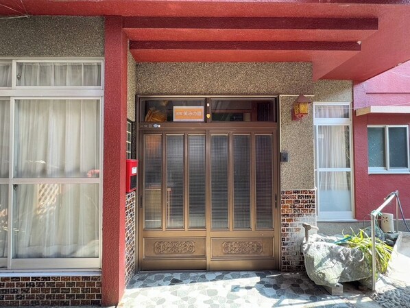A building entrance with a large wooden door, a tiled ground, and a small garden area with a rock and plants.