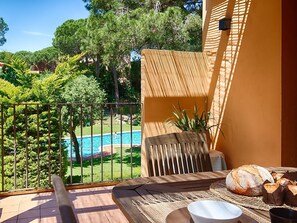 Un balcón con vista a una piscina, una silla de madera, una mesa con pan y un cuenco, y una planta en maceta.