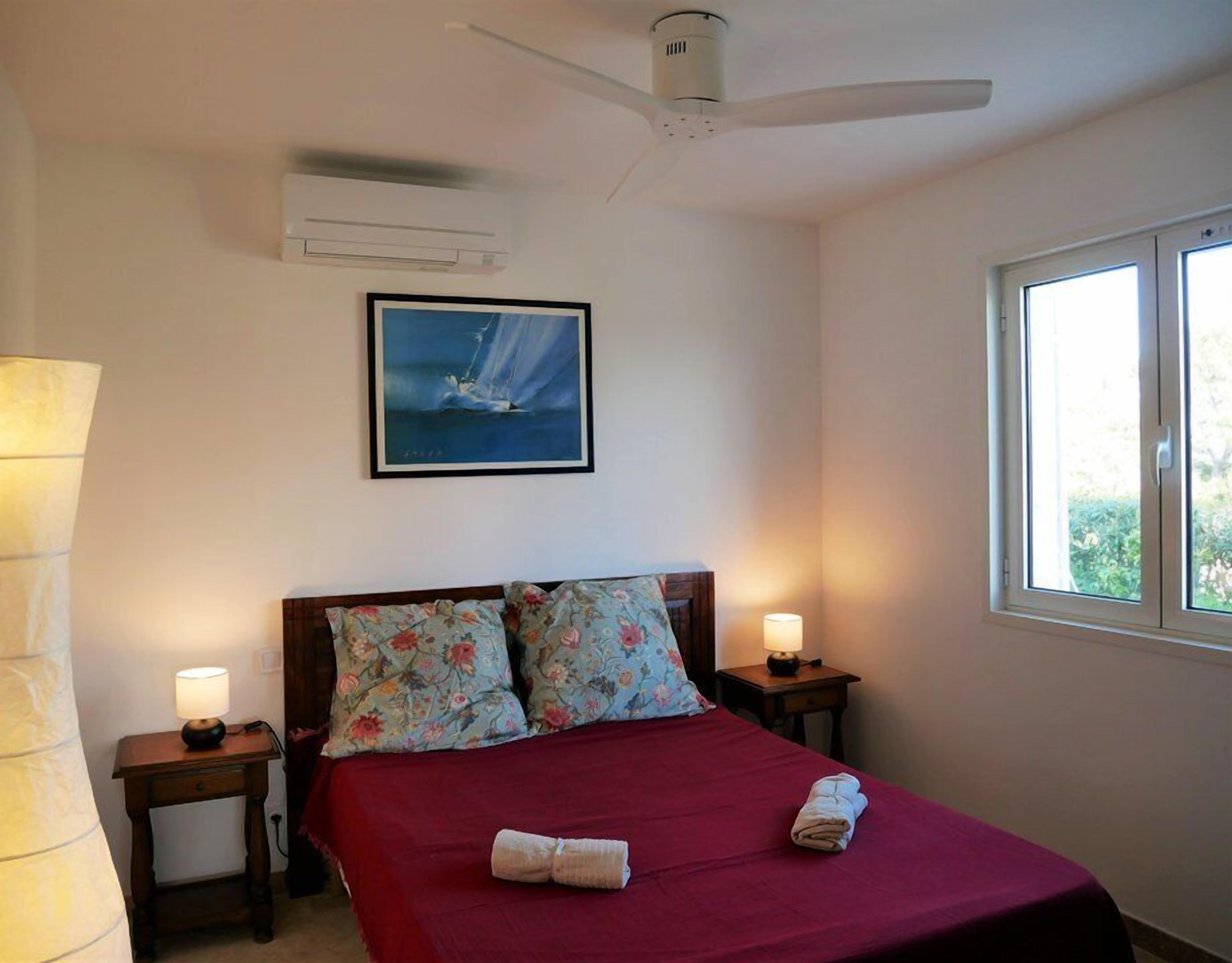 A bedroom with a bed, two bedside lamps, a ceiling fan, a painting, and a window.