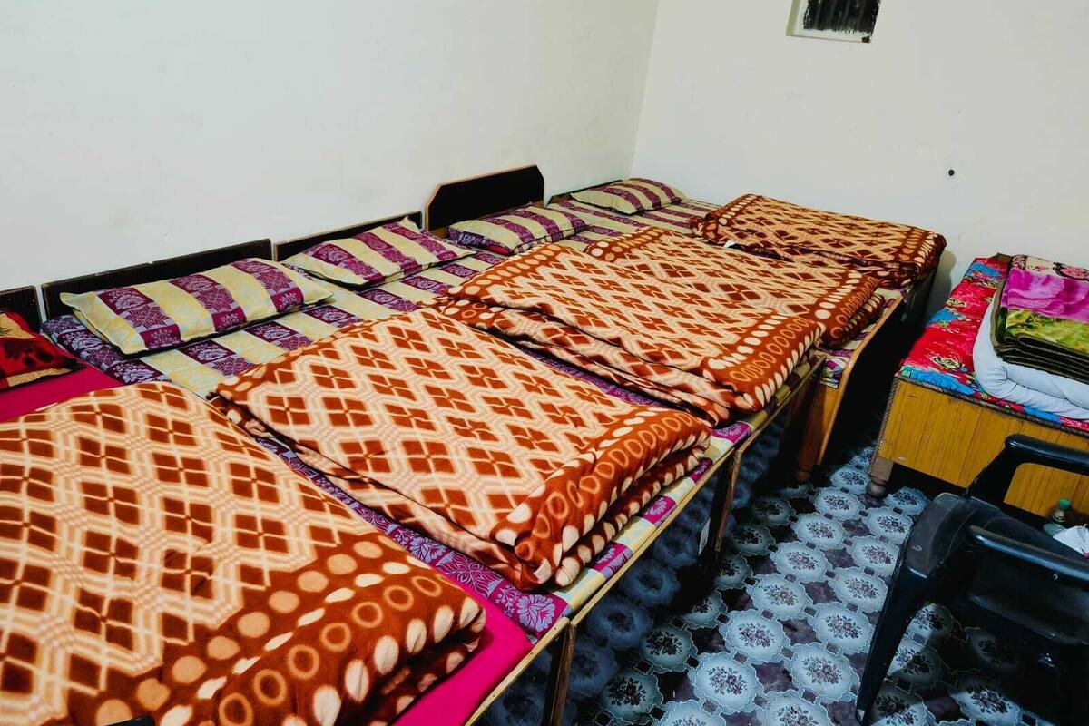 Une chambre de dortoir avec trois lits simples, chacun agrémenté de draps à motifs. On y trouve un bureau avec une chaise et une petite table avec une chaise. La chambre est aménagée de façon simple, avec une fenêtre et une petite grille d’aération sur le mur.