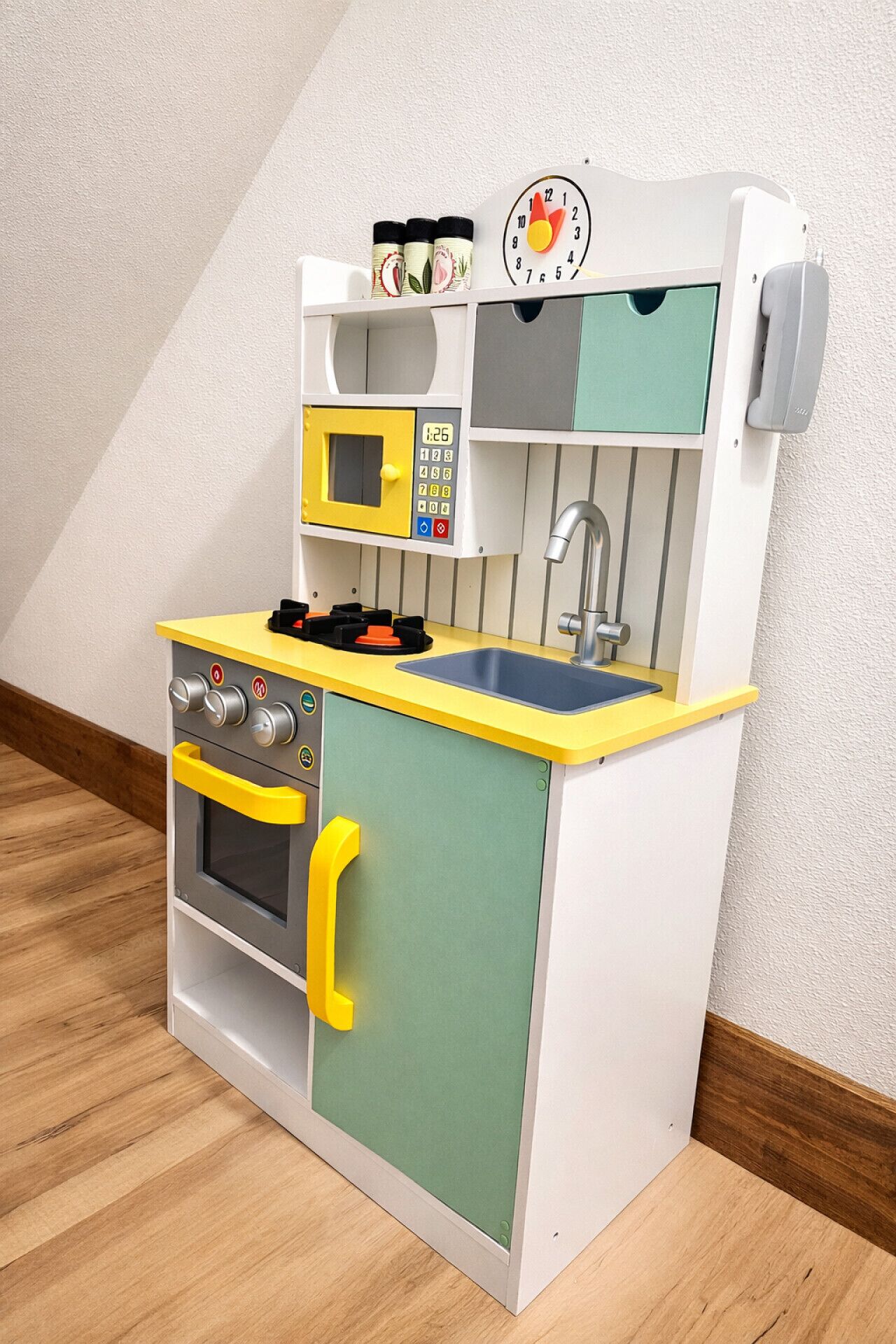 A children's play kitchen with a microwave, oven, sink, and a clock on the wall.