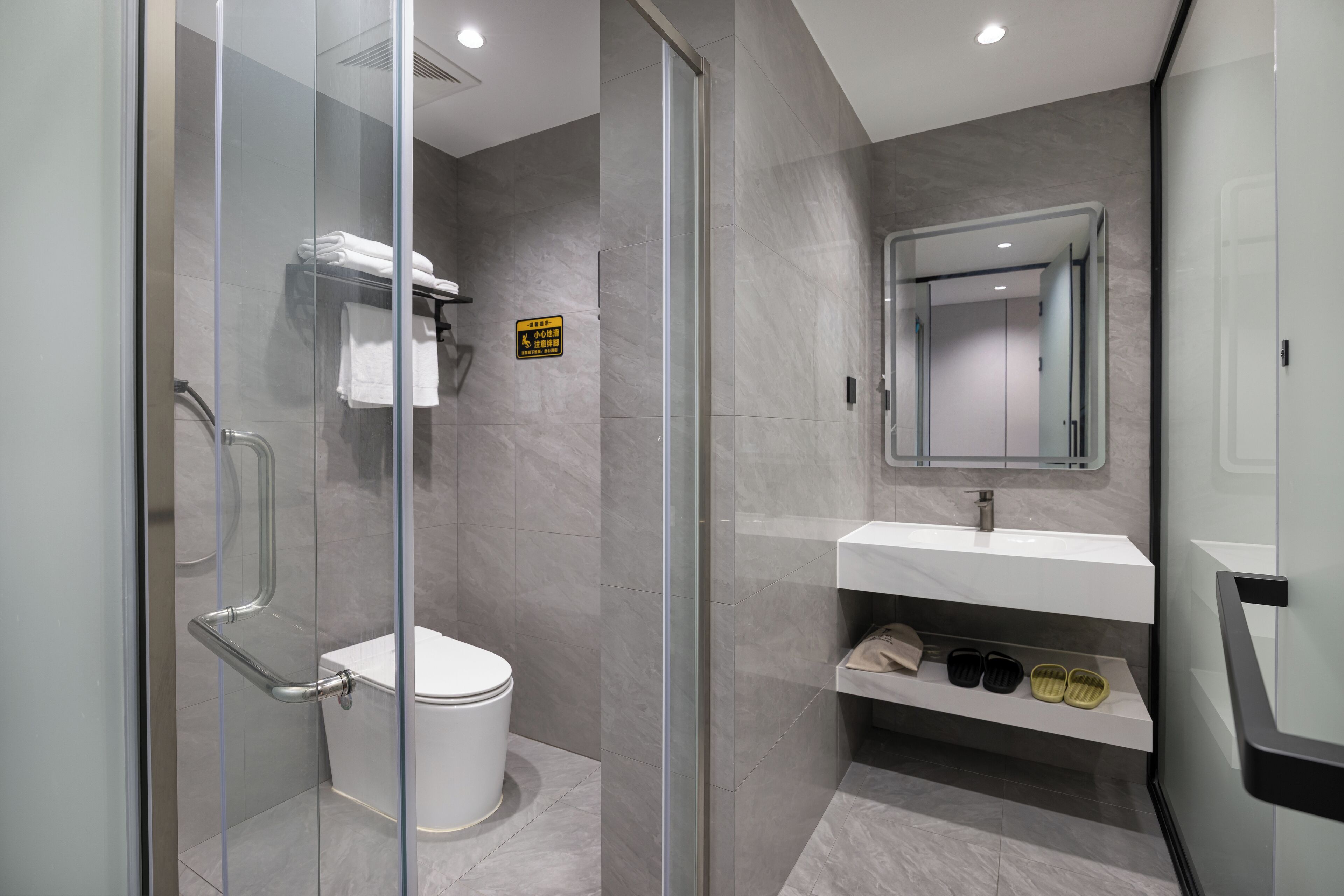 A modern bathroom with a glass-enclosed shower, a toilet, a sink with a mirror, and a shelf with slippers.