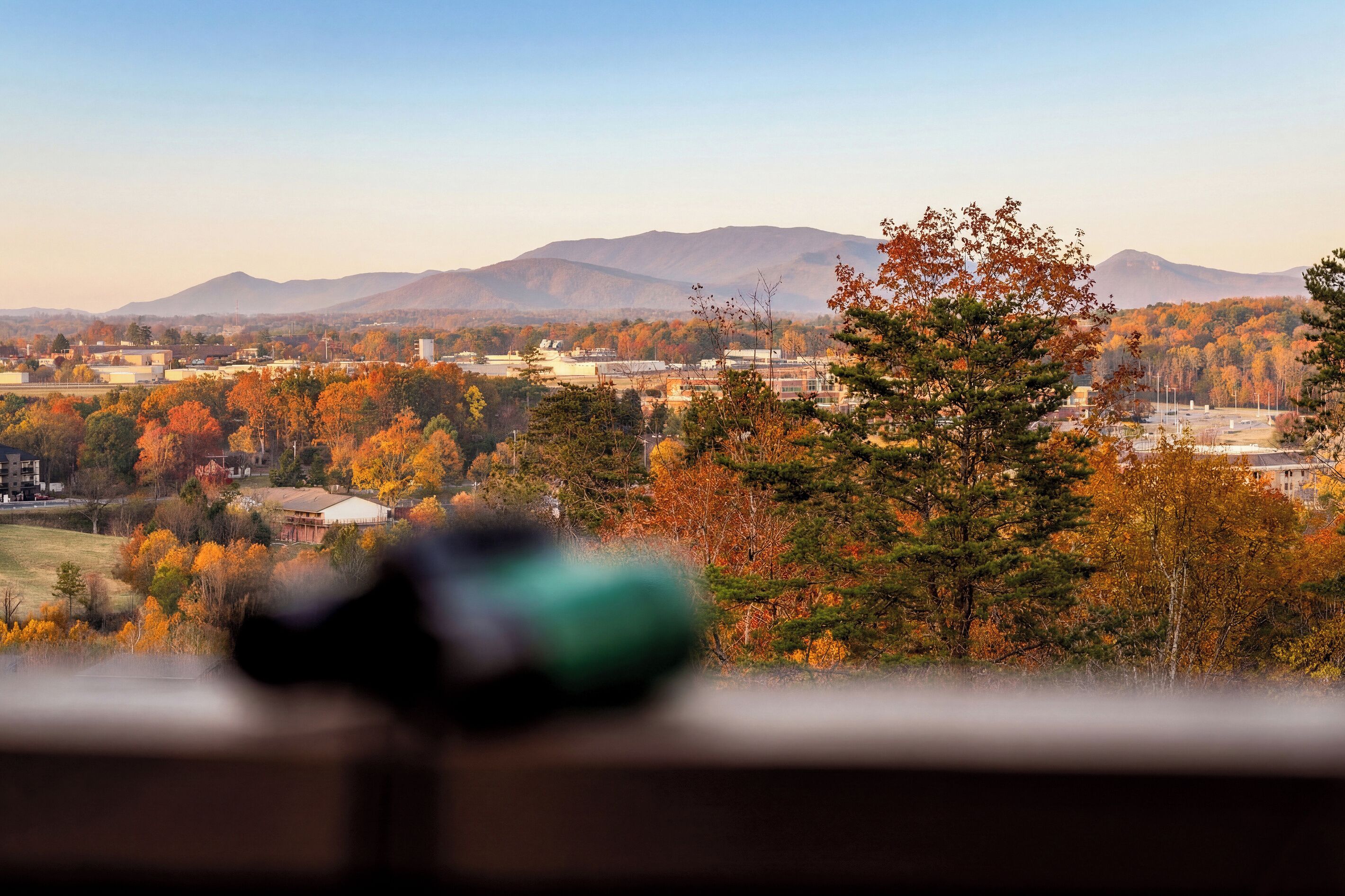 A panoramic view of a town with autumn foliage, mountains in the distance, and a blurred foreground object.