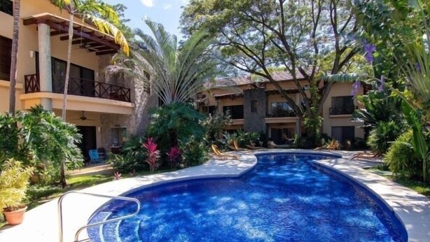 Una piscina rodeada de vegetación exuberante y un edificio de varios pisos con balcones.