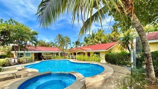 A swimming pool with a hot tub, surrounded by palm trees and a building with a red roof. - Cormoran A2 2 bed 2 Bath Condo In las Palmas (Playas del Coco)