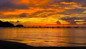 Atardecer sobre un mar tranquilo con barcos anclados, una playa de arena y un paisaje montañoso en la distancia.