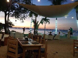 Een terras met tafels en stoelen, met uitzicht op een strand met boten en palmbomen.