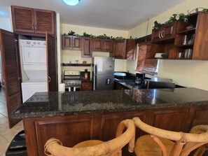 Una cocina con armarios de madera, encimera de granito, refrigerador, estufa y dos sillas de mimbre.
