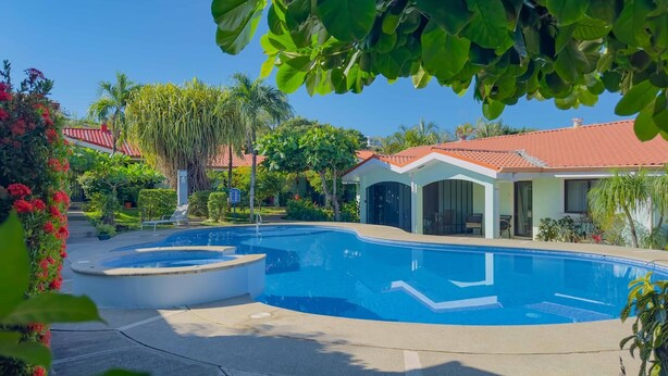 A swimming pool with a curved edge, surrounded by a concrete deck. There is a covered seating area with a red-tiled roof and glass doors. - Green Life #3 | Perfect Beachside Living (Playas del Coco)