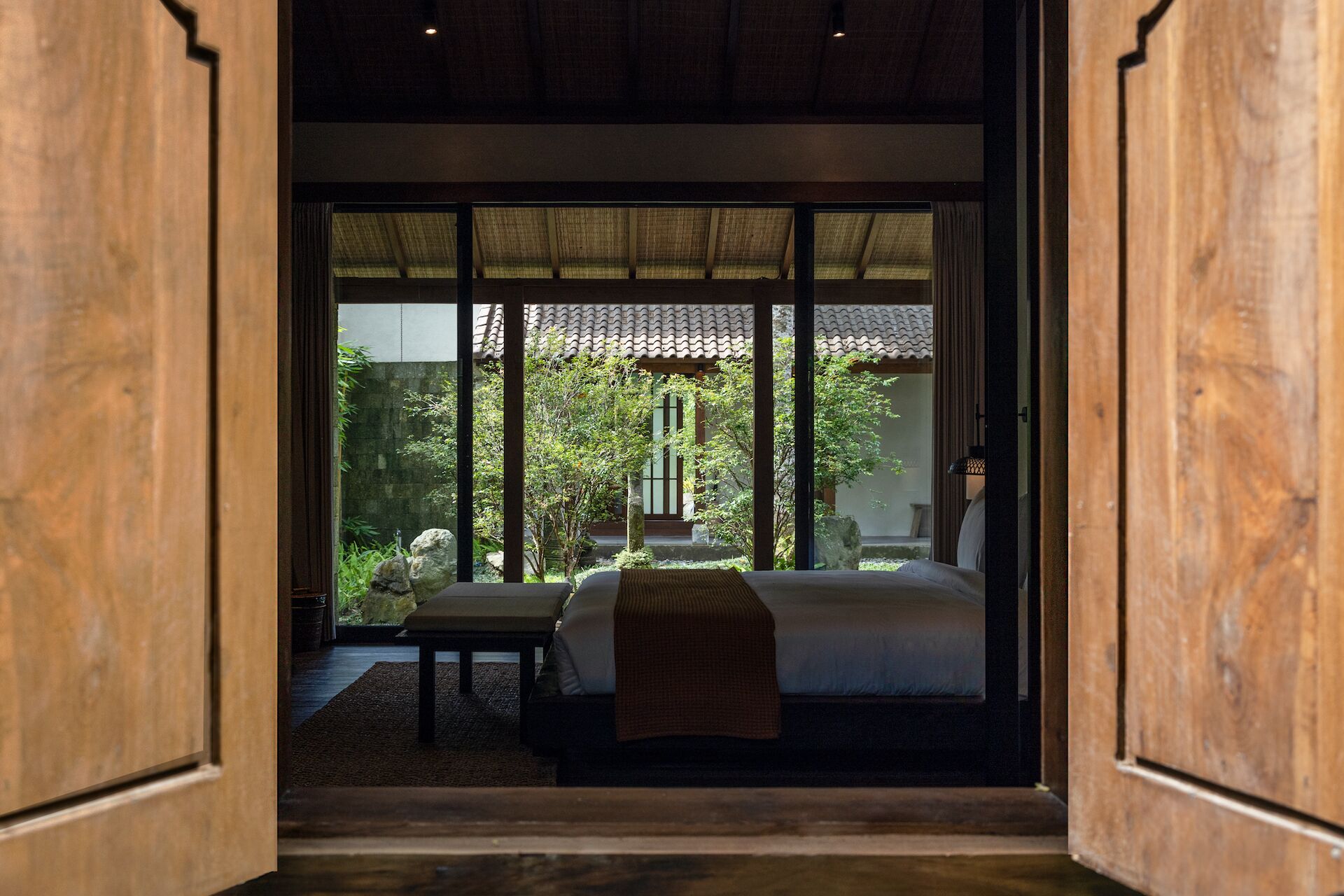 Un dormitorio tradicional japonés con suelo de tatami, cama baja y vistas a un jardín con farol de piedra y un árbol.
