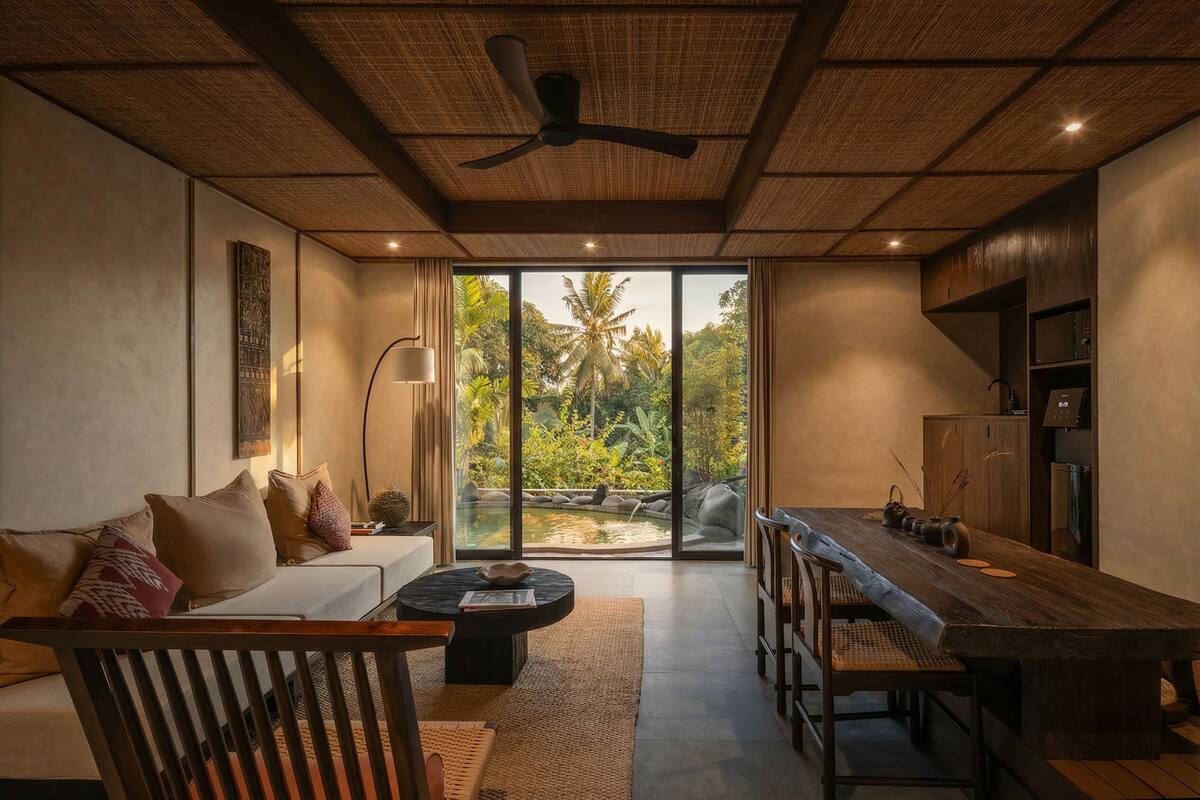 A modern living room with a sofa, coffee table, dining table, and chairs. There is a ceiling fan, a large window with a view of greenery, and a kitchen area with cabinets and appliances.