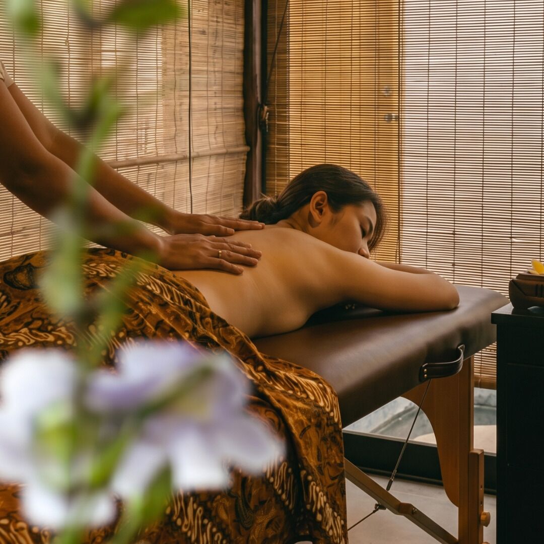 Une séance de massage dans une pièce équipée d’une table de massage en bois, d’une fenêtre avec des stores et d’une petite table sur laquelle se trouve un bol.