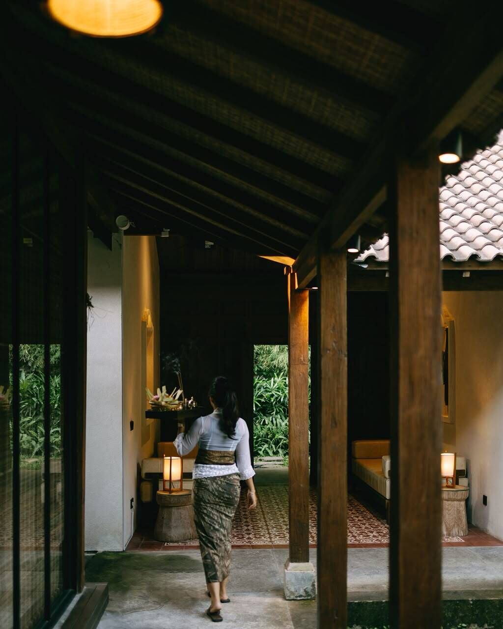 A covered walkway with wooden pillars, a person walking, and a view of greenery outside.