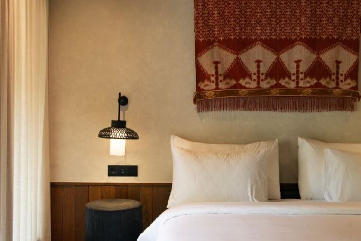 Habitación de hotel con cama, lámpara de pared, taburete redondo y un tapiz estampado.