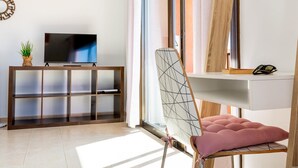 Una sala de estar moderna con un televisor de pantalla plana sobre un mueble de madera, una silla de vidrio con un cojín rosa y un estante blanco con un espejo y gafas de sol.
