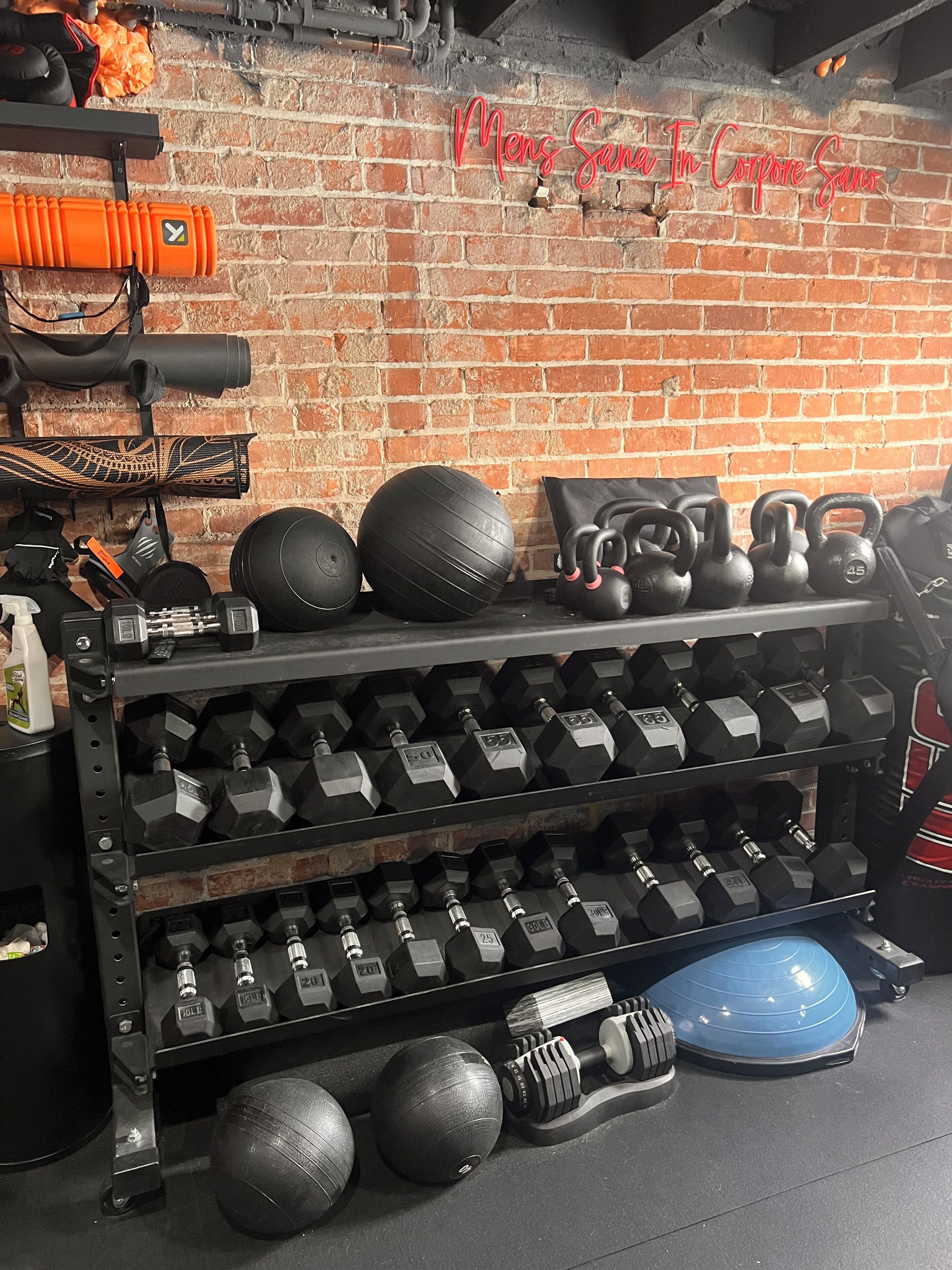 A gym with a brick wall, various exercise equipment, and a sign that reads "Mens Sana In Corpore Sanus".
