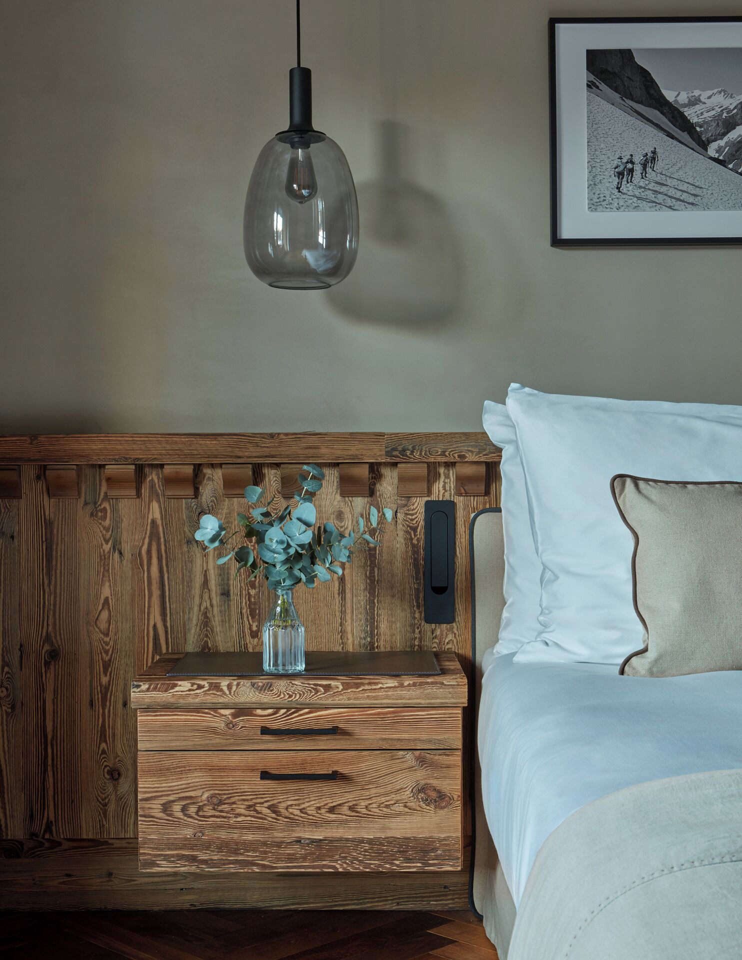 Una mesita de noche de madera con un florero, una cama con ropa de cama blanca y un cuadro en la pared.