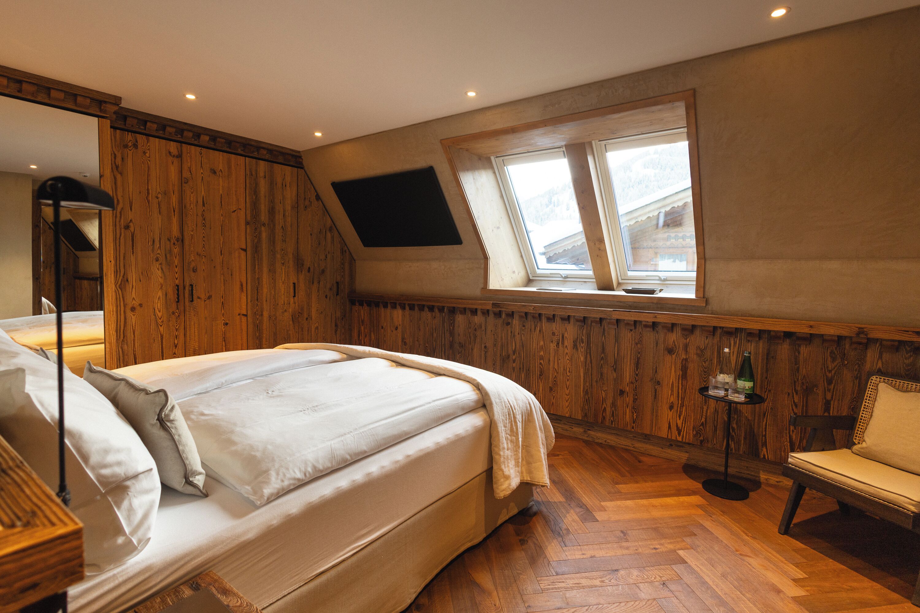 A bedroom with a wooden ceiling, a bed with white linens, a window with a view, a small table with a bottle, and a chair.