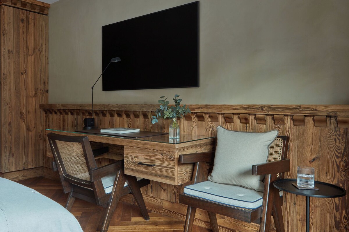 Habitación con paneles de madera, un escritorio, dos sillas, una mesita con un vaso y un televisor de pantalla plana montado en la pared.