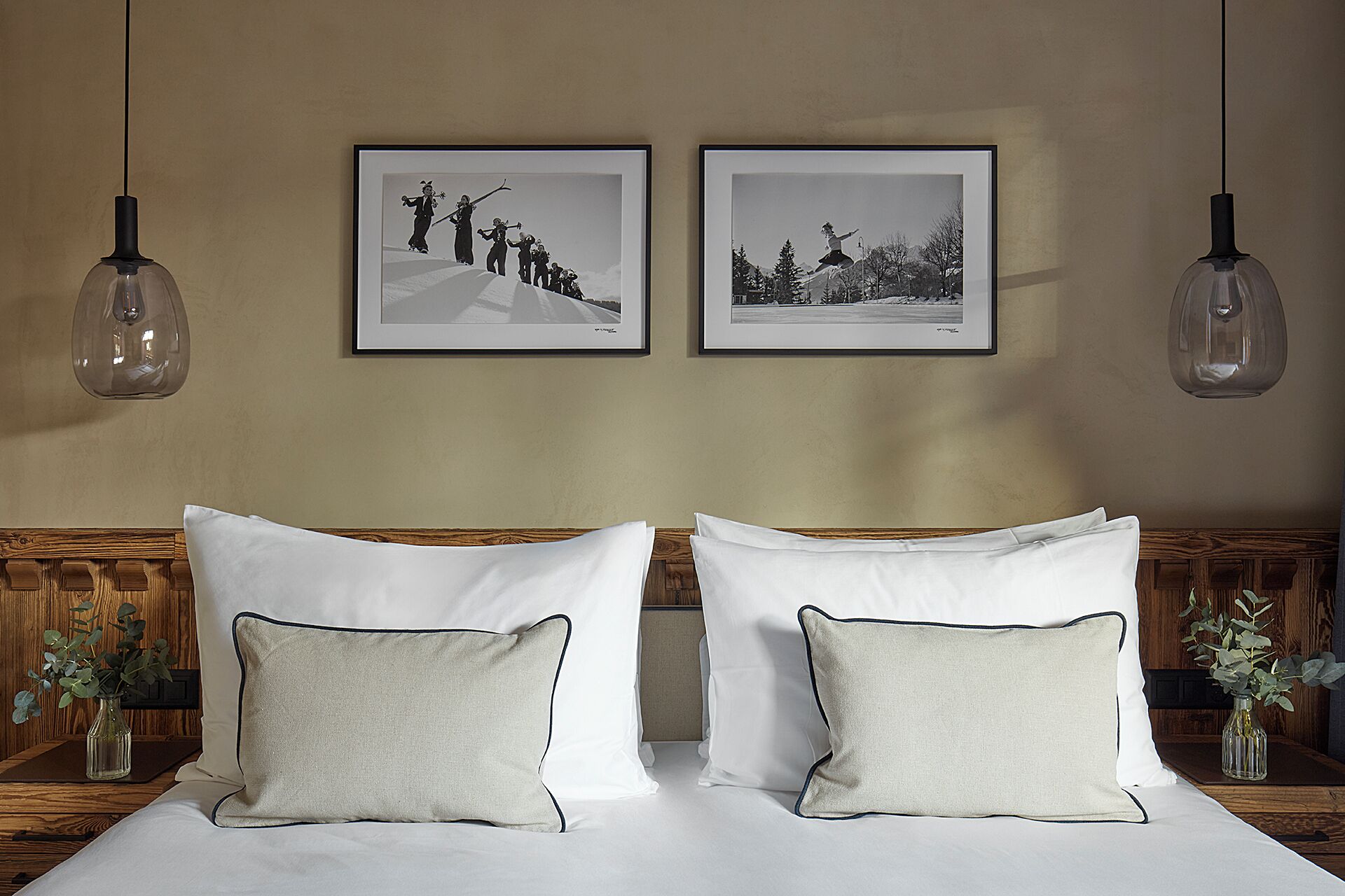 Una cama doble bien tendida con sábanas blancas y almohadas a juego, dos fotografías enmarcadas en la pared y lámparas colgantes.