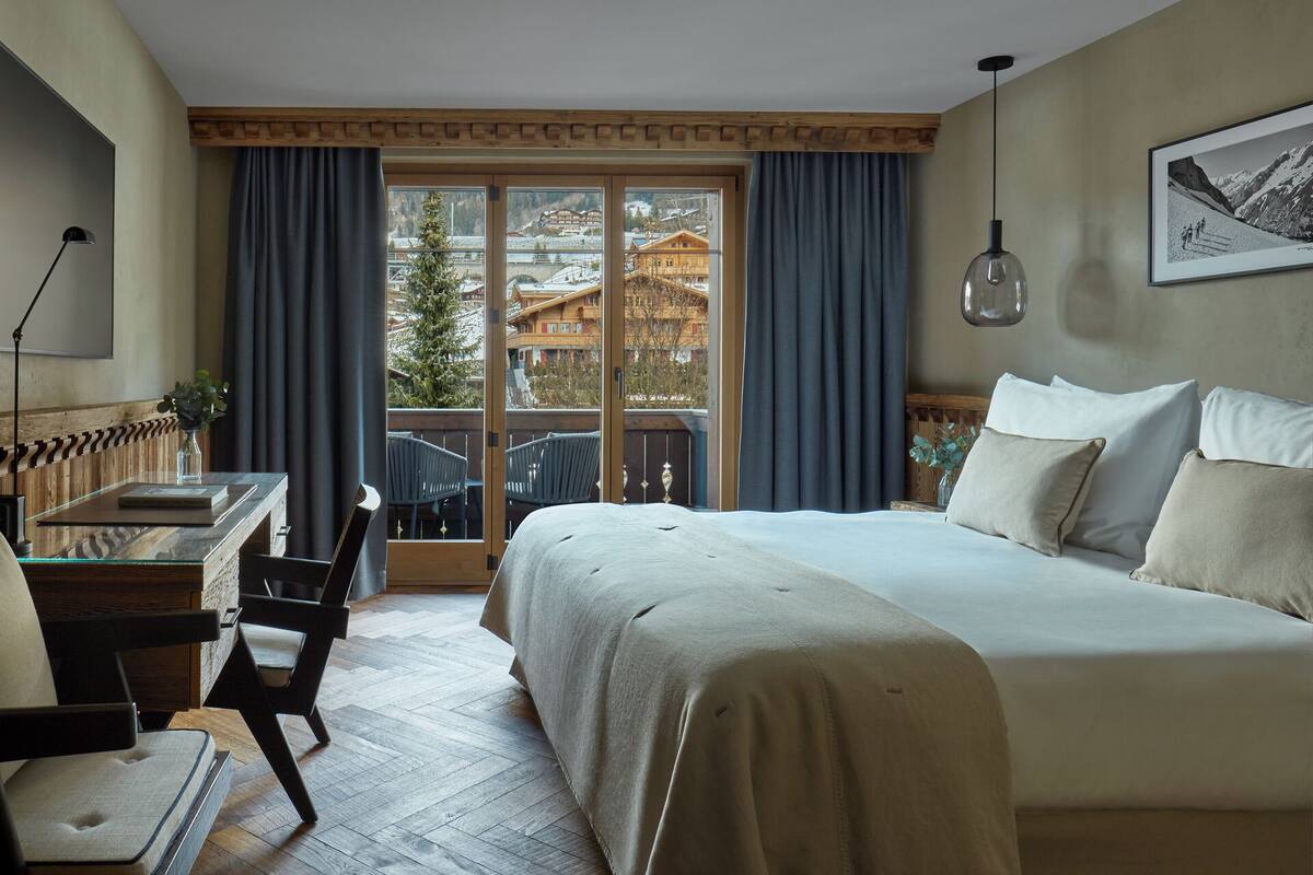 Una camera d'albergo con un letto, una scrivania, una sedia e vista sugli edifici attraverso ampie finestre.