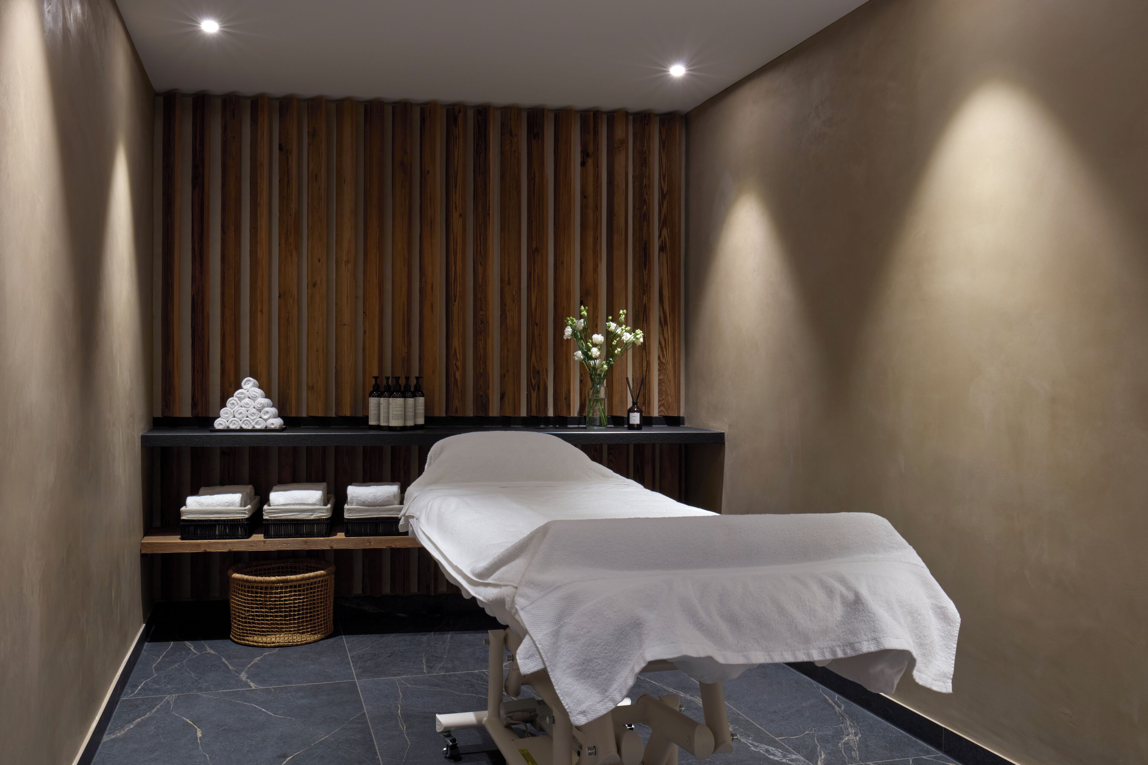 Een spa-ruimte met een massagetafel, een wand van houten panelen, planken met handdoeken en flessen, en een rieten mand.