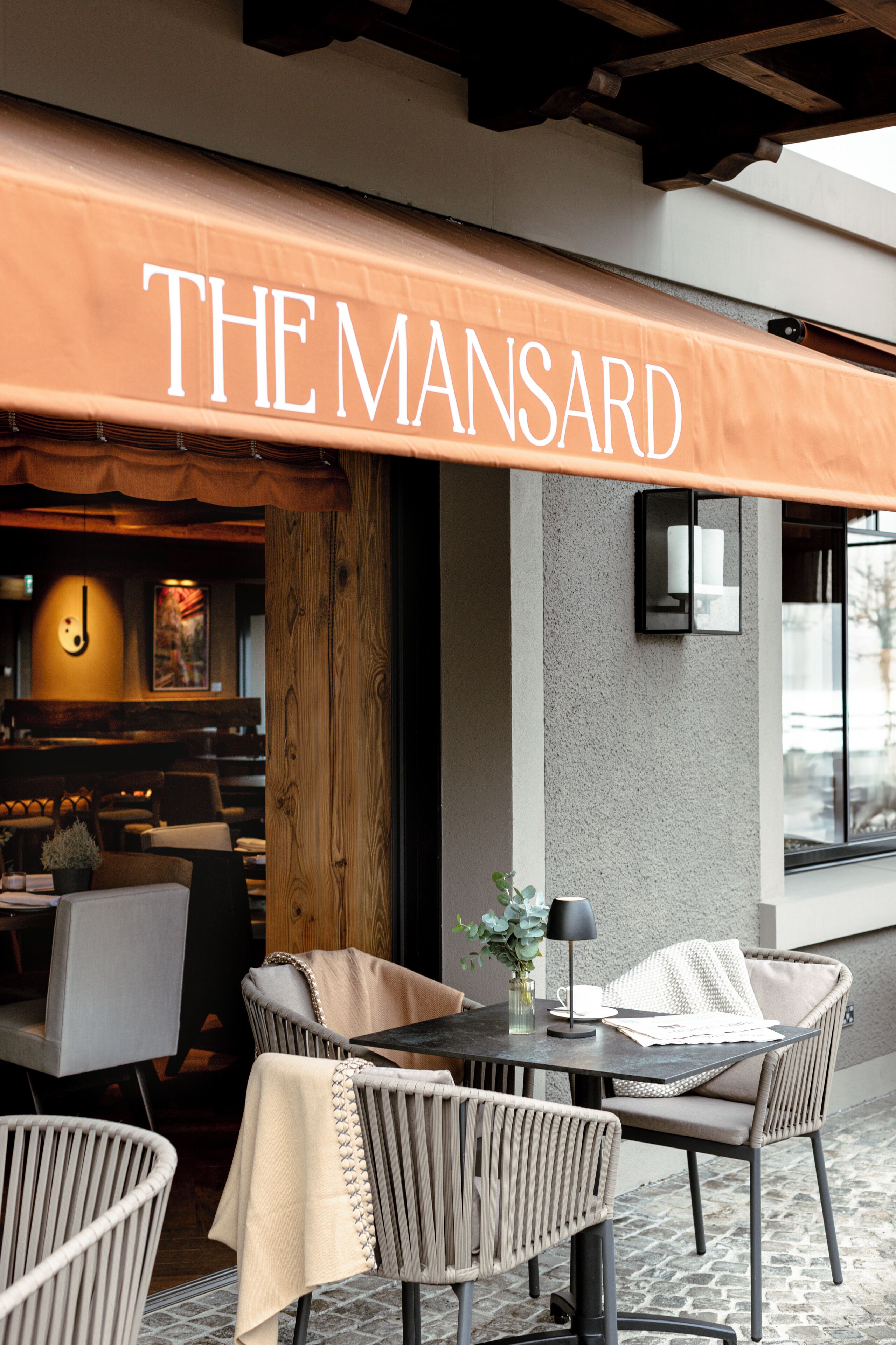 An outdoor seating area with wicker chairs and a table, a wooden column, and a sign that reads "THE MANSARD".