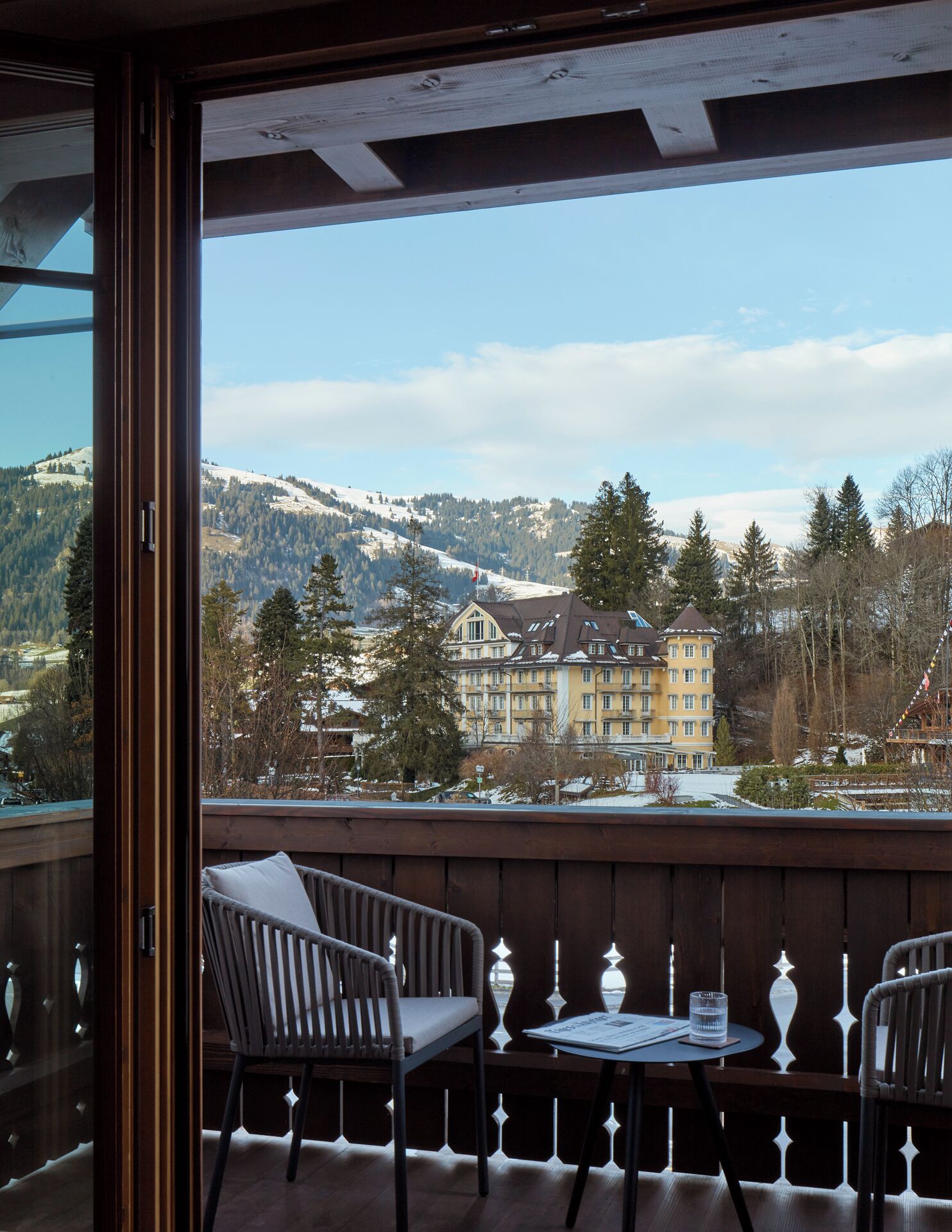 A balcony with two chairs and a small table, overlooking a mountain and a building with multiple windows.