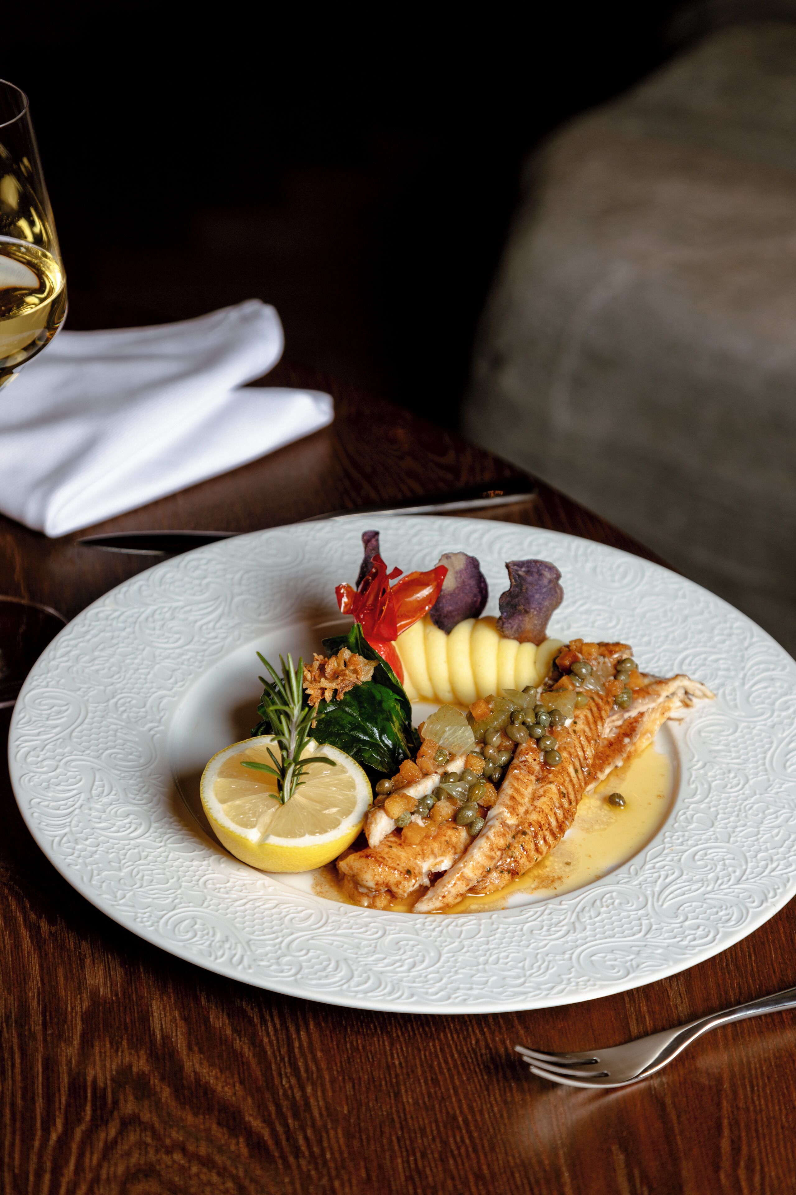 A plate of fish with lemon and herbs, accompanied by a glass of white wine and a napkin on a wooden table.