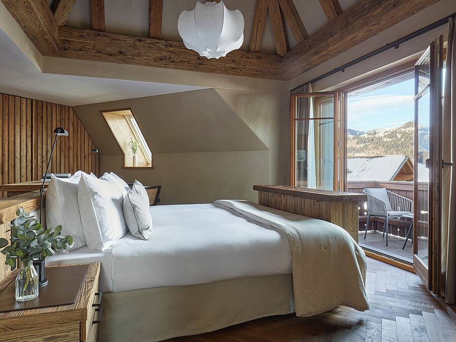 A bedroom with a large bed, wooden beams, a skylight, a view of the outdoors, and a balcony with chairs.