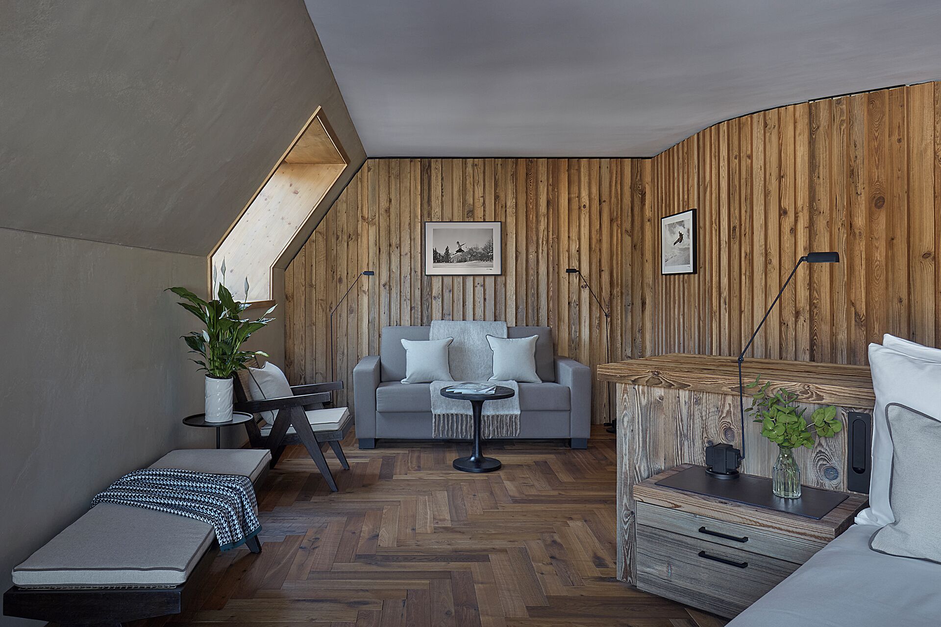 Ein modernes Schlafzimmer im Loft-Stil mit schrägem Dach, Holzwandverkleidung, einem grauen Sofa, einem kleinen Tisch, einer Bank und einem Nachttisch mit Lampe.