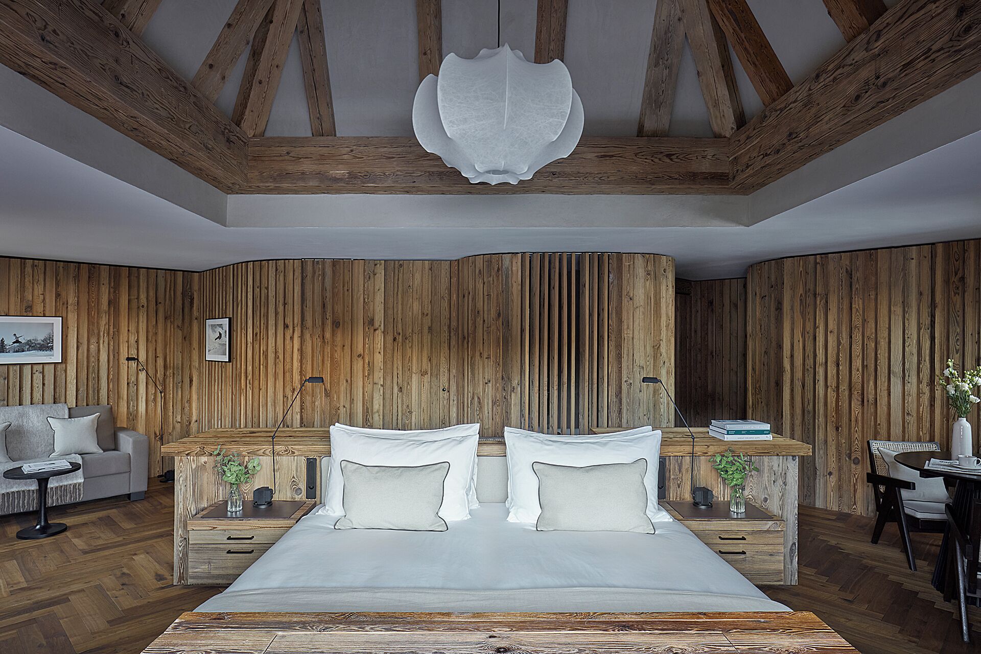 A bedroom with a wooden ceiling, a bed with white bedding, a wooden nightstand, a desk, and a chair.