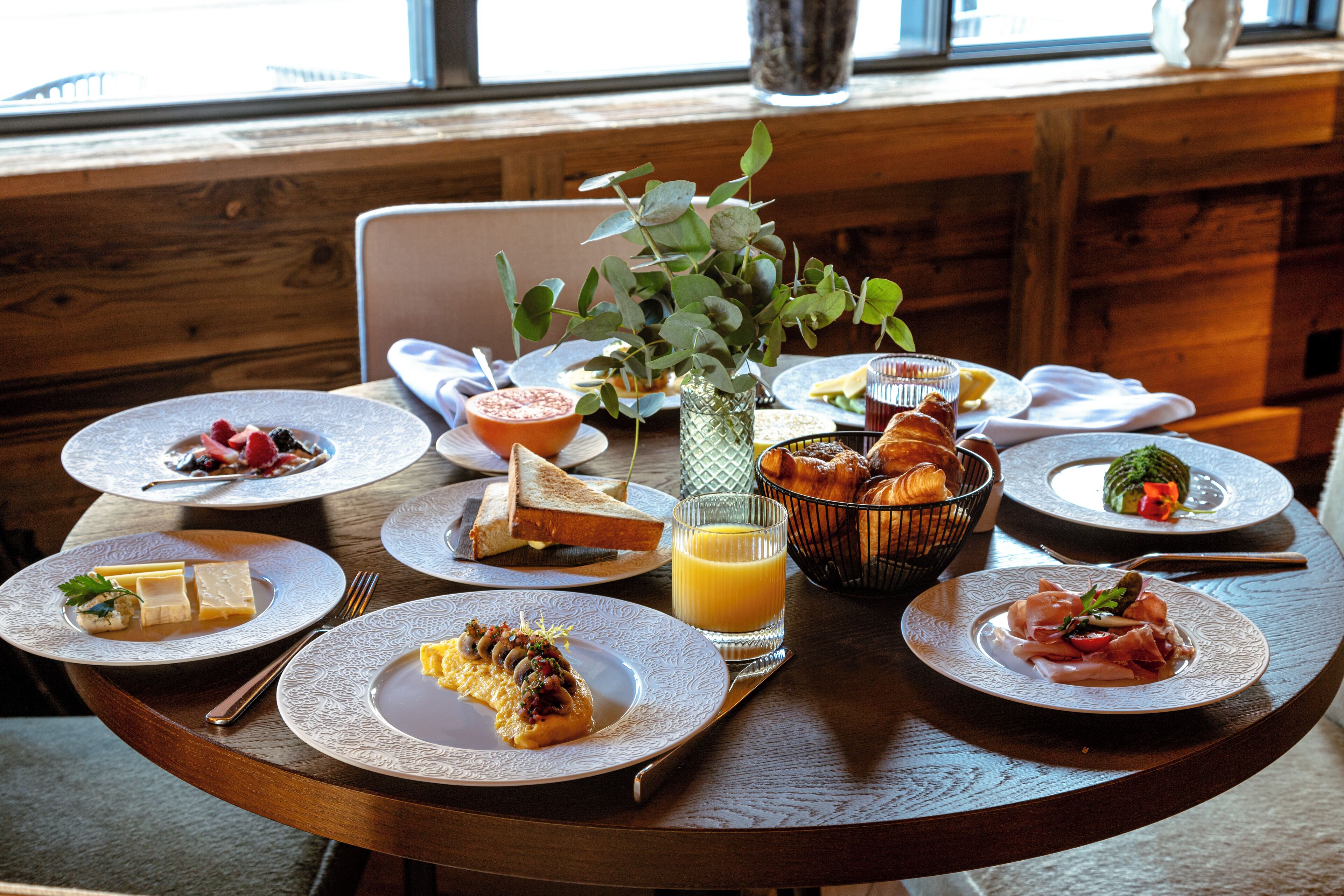 A breakfast setting with multiple plates of food, a glass of orange juice, and a vase with greenery.