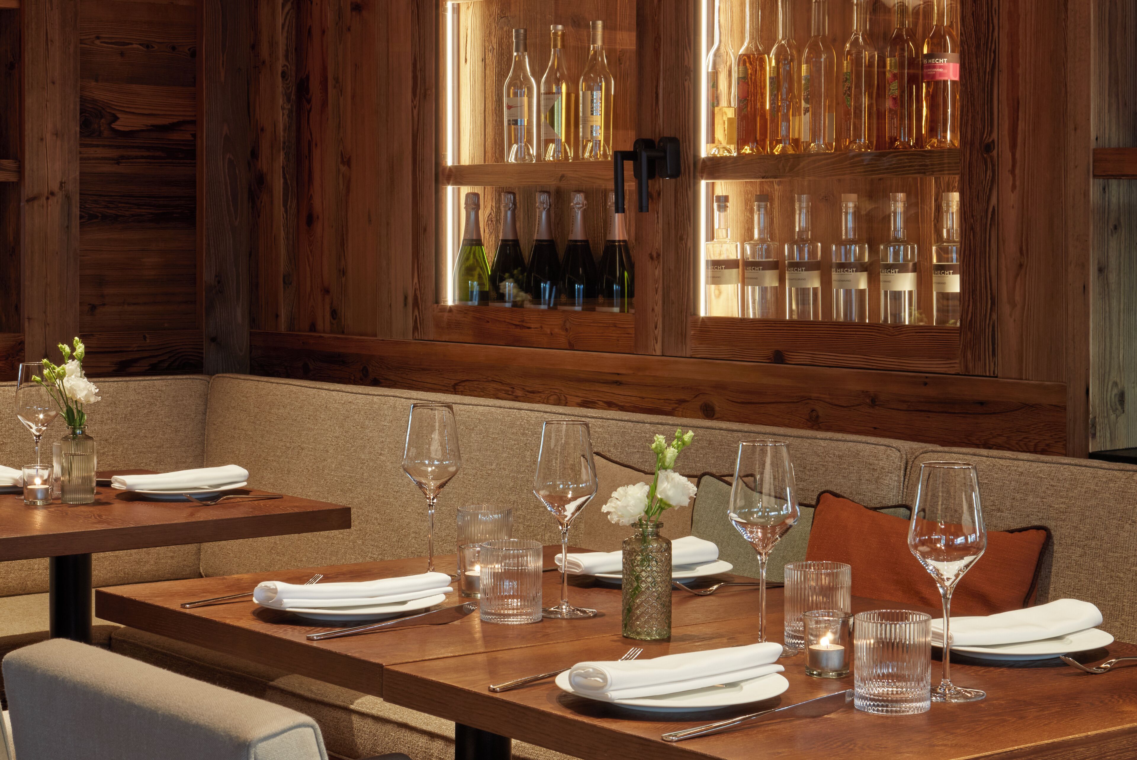 A restaurant interior with wooden walls, a bar area with bottles, and tables set for dining with wine glasses and napkins.