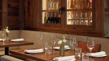 A restaurant interior with wooden walls, a bar area with bottles, and tables set for dining with wine glasses and napkins.