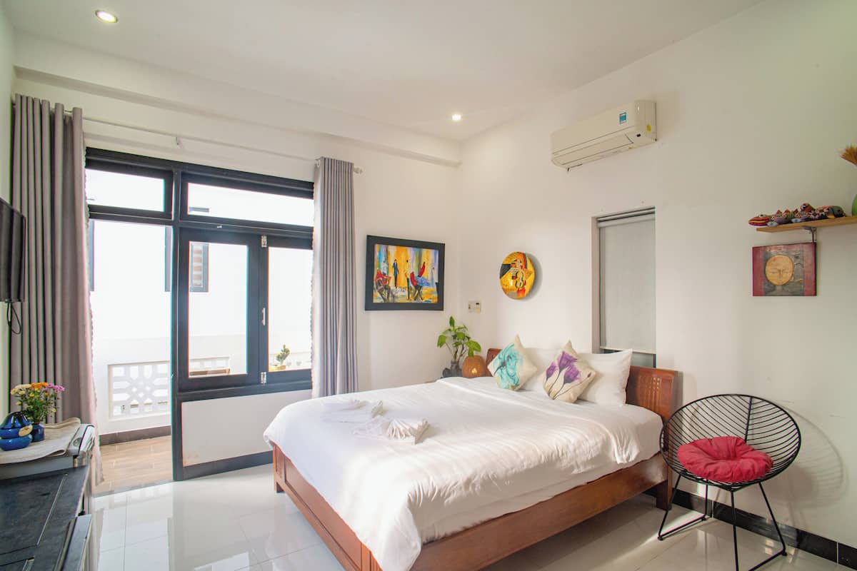 A modern hotel room with a large bed, a chair, a window with curtains, and artwork on the walls.