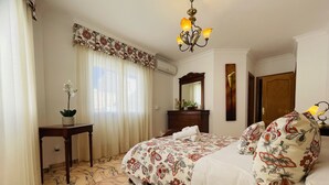A hotel room with a bed, a desk, a chair, a mirror, a chandelier, and a window with curtains.