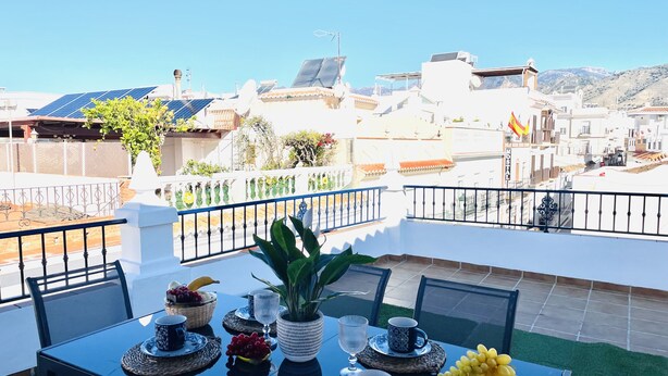 A rooftop terrace with a table set for two, featuring a potted plant, a bowl of grapes, and a basket of bread.