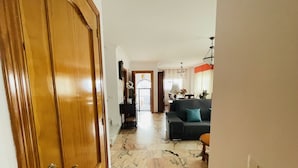 A hallway with a wooden door, a marble floor, a dark-colored sofa with a teal pillow, and a dining area in the background.