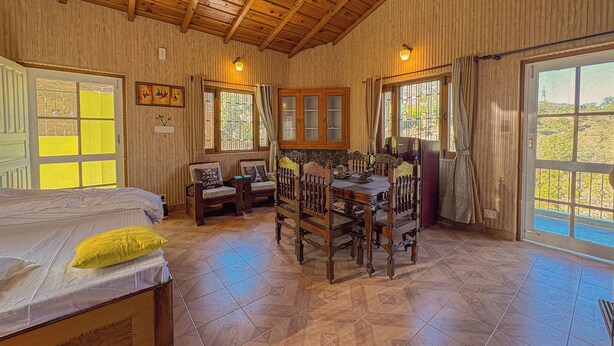 Una habitación acogedora con techo de madera, una mesa de comedor con sillas, una cama con almohada amarilla y ventanas amplias con cortinas.