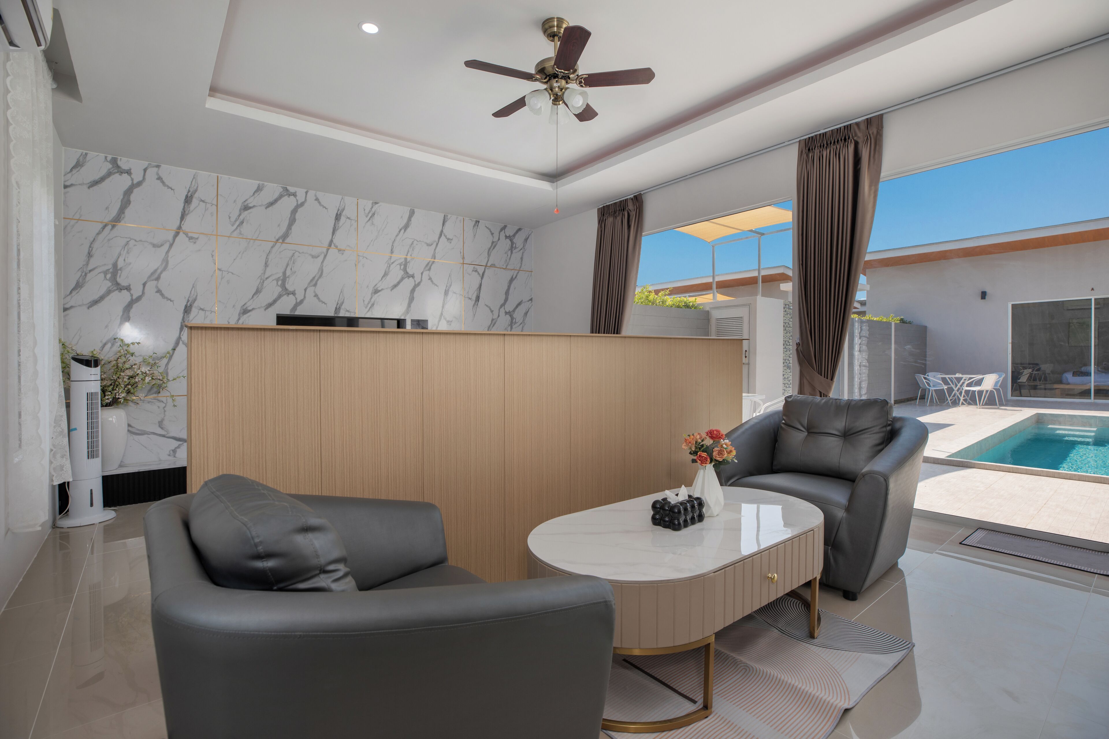 A modern living room with a ceiling fan, a marble wall, a poolside area with a table and chairs, and a view of the pool.